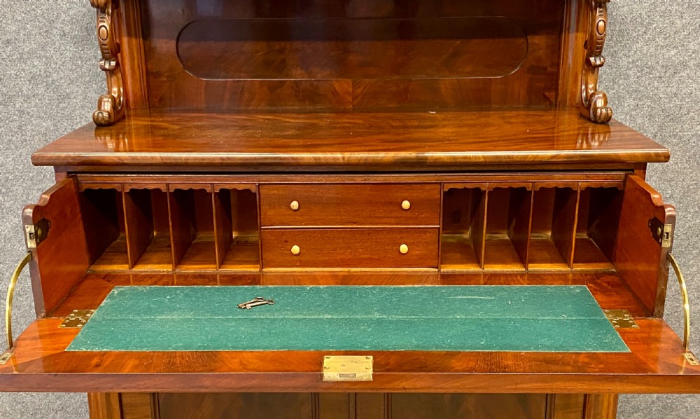 A Victorian flame mahogany secretaire chiffonier featuring an ornate carved cresting top shelf, drop-front desk compartment with green writing surface, and a two-door cupboard base. View VI