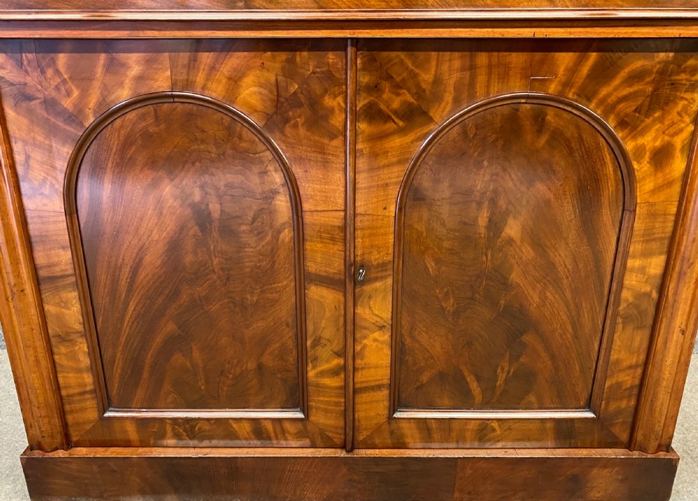 A Victorian flame mahogany secretaire chiffonier featuring an ornate carved cresting top shelf, drop-front desk compartment with green writing surface, and a two-door cupboard base. View VII