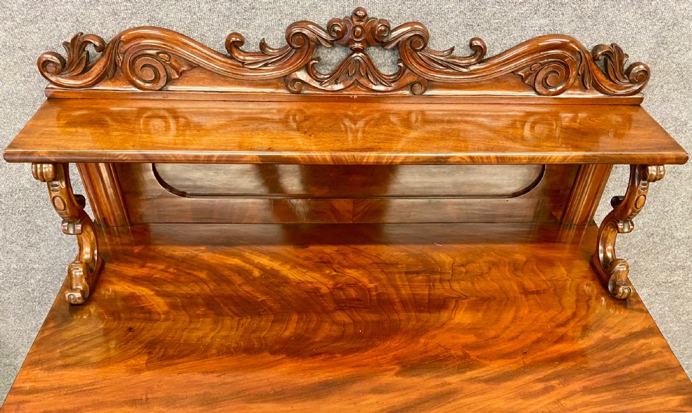 A Victorian flame mahogany secretaire chiffonier featuring an ornate carved cresting top shelf, drop-front desk compartment with green writing surface, and a two-door cupboard base. View IX