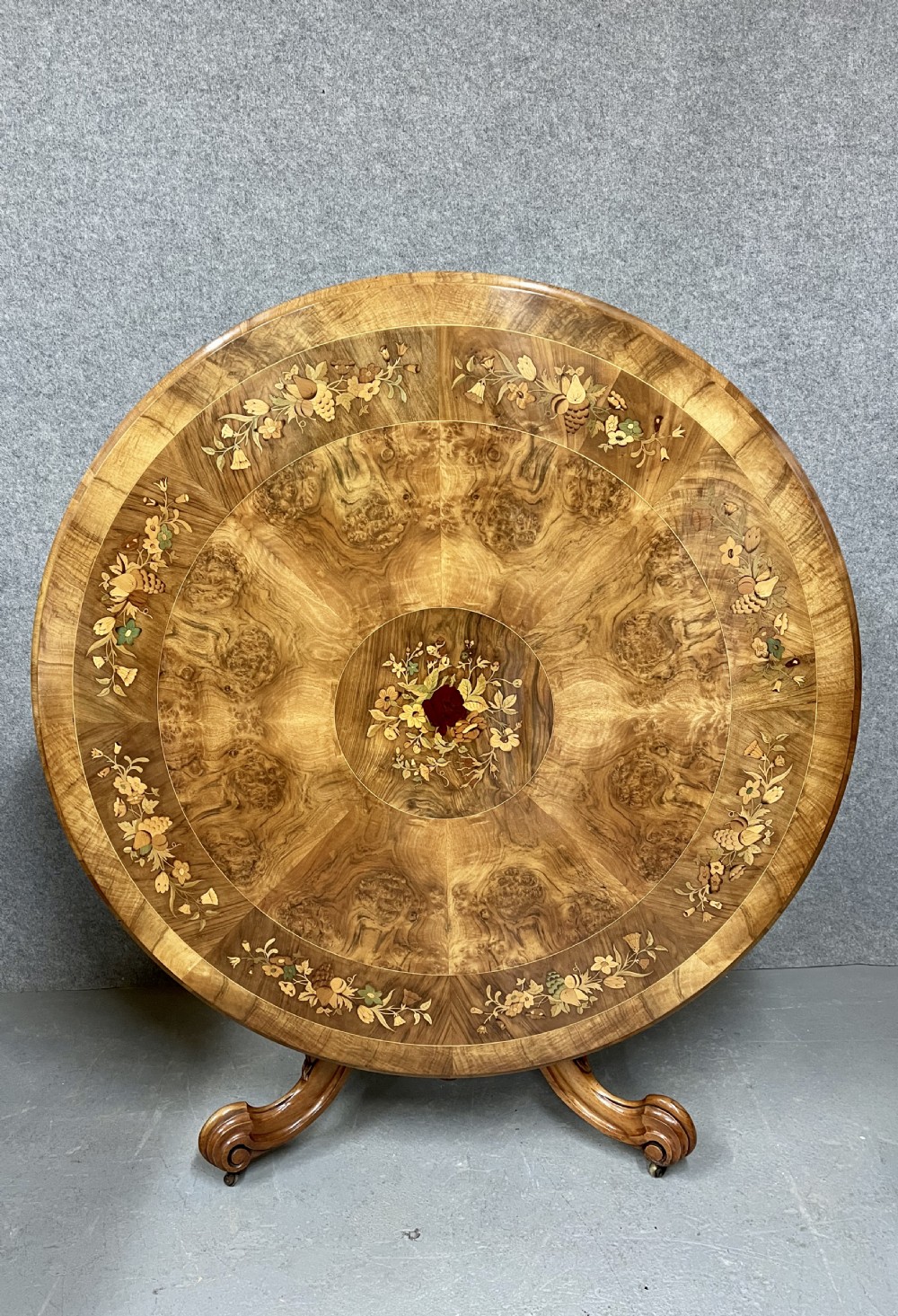 A circular Victorian burr walnut centre table featuring floral marquetry inlay on the top surface and supported by a carved pedestal base.