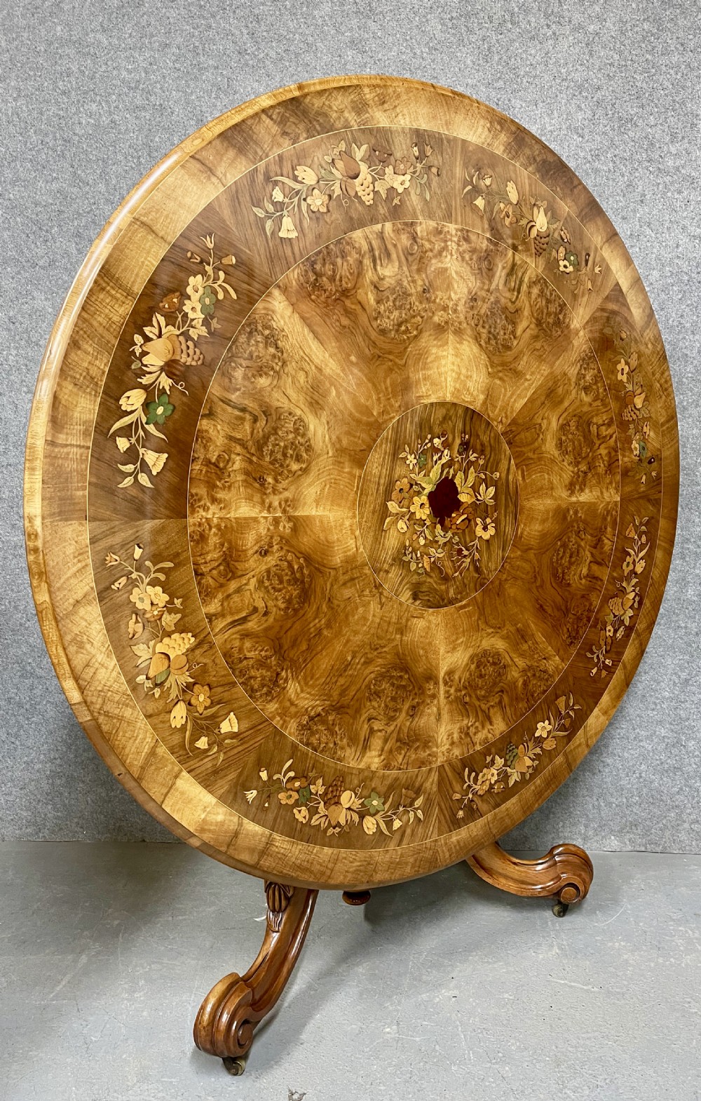 A circular Victorian burr walnut centre table featuring floral marquetry inlay on the top surface and supported by a carved pedestal base. View II