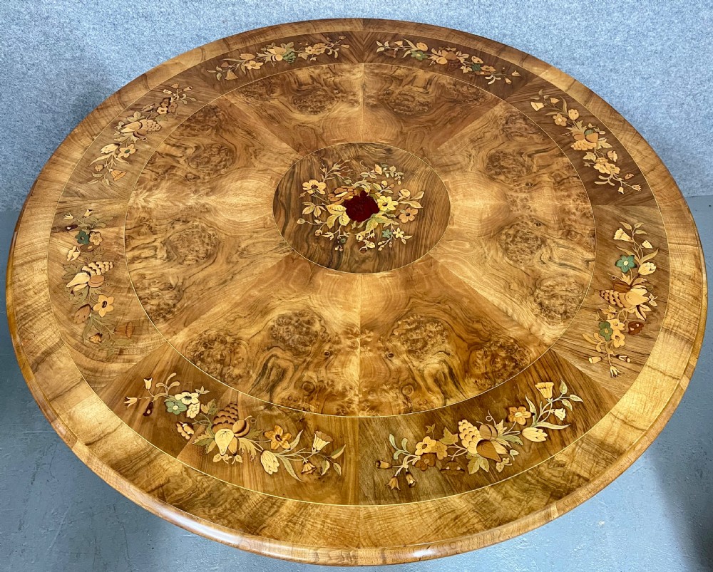 A circular Victorian burr walnut centre table featuring floral marquetry inlay on the top surface and supported by a carved pedestal base. View X