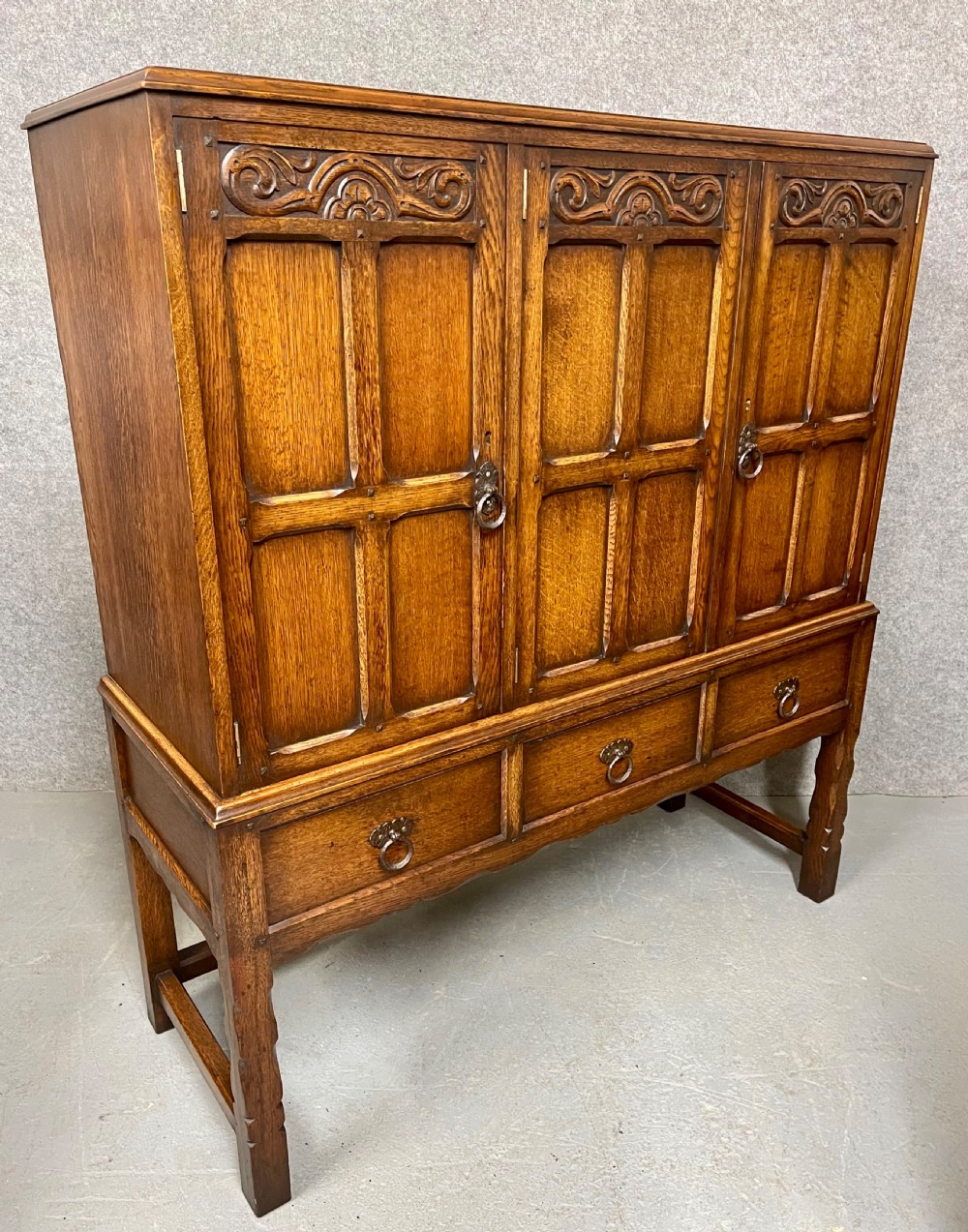 A warm brown oak cocktail cabinet featuring three carved panel doors above three drawers, set on a sturdy base with square legs.