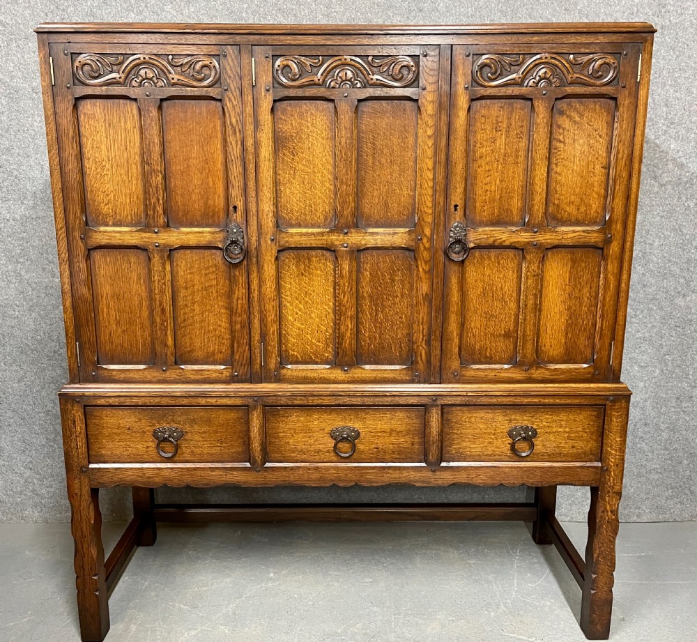 A warm brown oak cocktail cabinet featuring three carved panel doors above three drawers, set on a sturdy base with square legs. View II