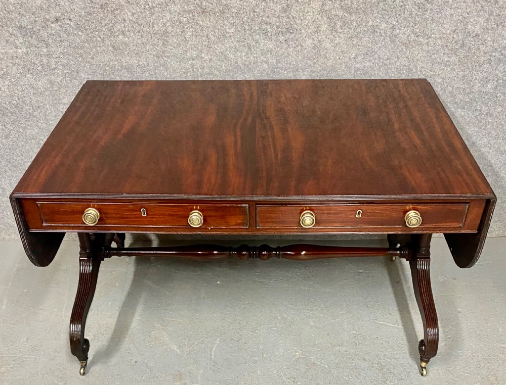A Regency figured mahogany sofa table featuring two front drawers with brass knobs, drop-down side leaves, a turned stretcher, and reeded sabre legs with brass castors.