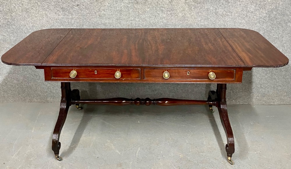A Regency figured mahogany sofa table featuring two front drawers with brass knobs, drop-down side leaves, a turned stretcher, and reeded sabre legs with brass castors. View II