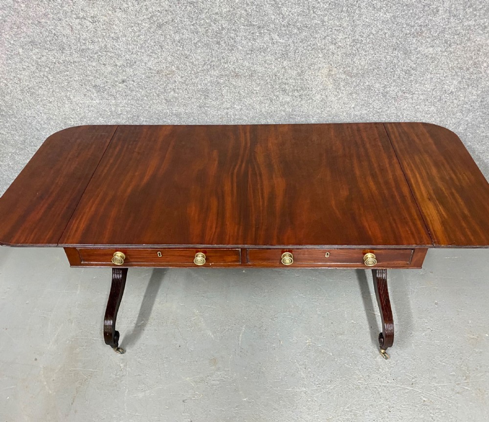 A Regency figured mahogany sofa table featuring two front drawers with brass knobs, drop-down side leaves, a turned stretcher, and reeded sabre legs with brass castors. View III
