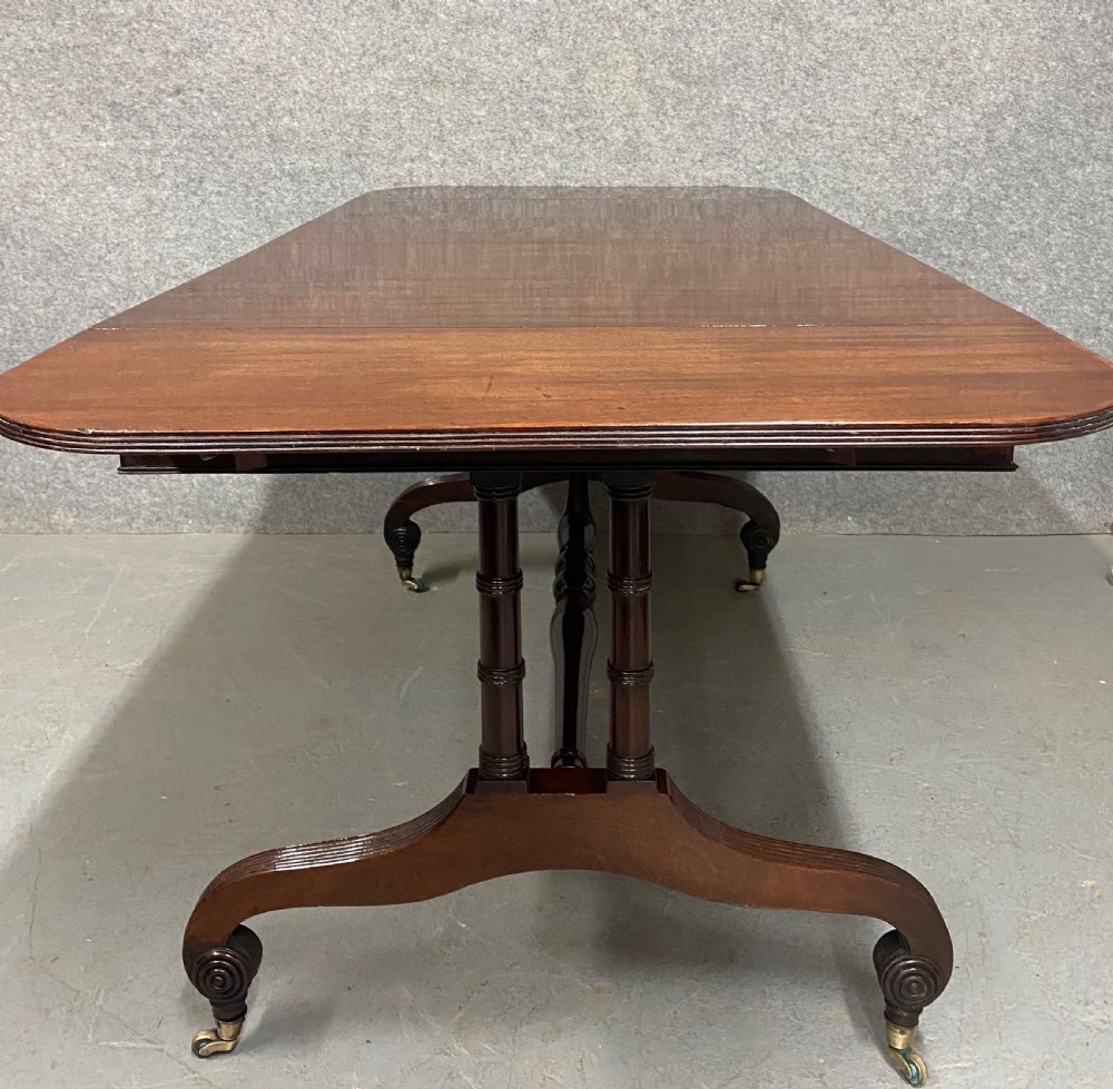 A Regency figured mahogany sofa table featuring two front drawers with brass knobs, drop-down side leaves, a turned stretcher, and reeded sabre legs with brass castors. View IV