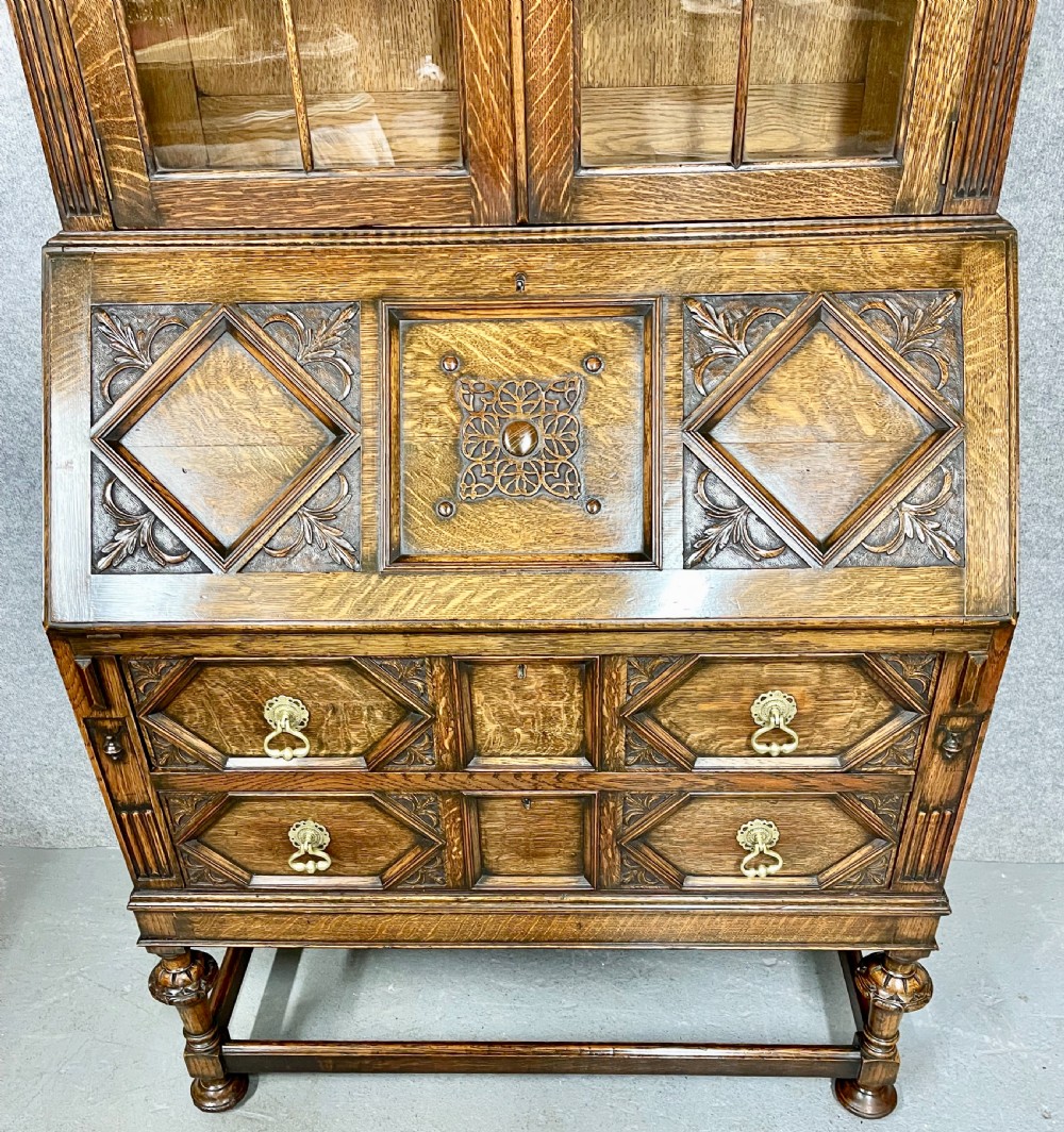 A carved oak bureau bookcase featuring an upper glazed two-door cabinet, a fall-front writing slope with geometric panels, and drawers set upon turned legs. View VII