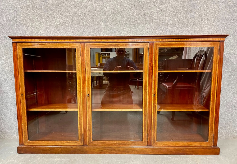 A wide, low Edwardian mahogany bookcase featuring three glass doors accented with satinwood crossbanding and internal wooden shelves on a plinth base. View IX