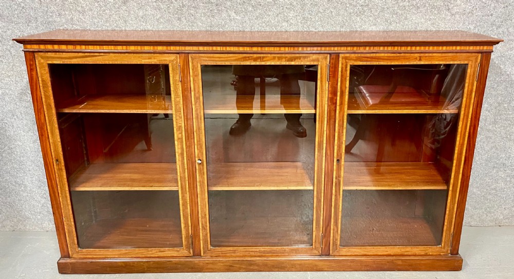 A wide, low Edwardian mahogany bookcase featuring three glass doors accented with satinwood crossbanding and internal wooden shelves on a plinth base. View II