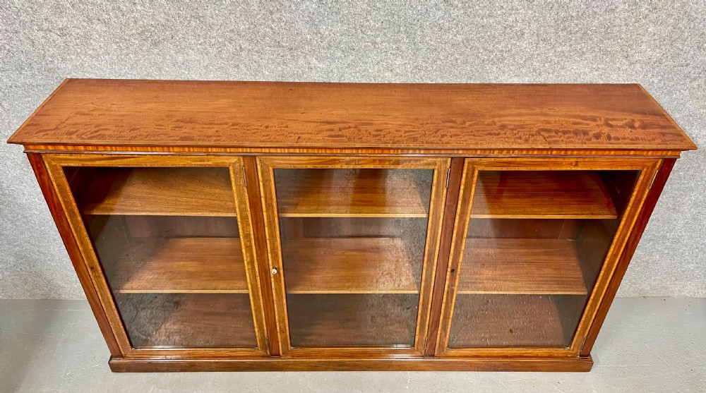 A wide, low Edwardian mahogany bookcase featuring three glass doors accented with satinwood crossbanding and internal wooden shelves on a plinth base. View III
