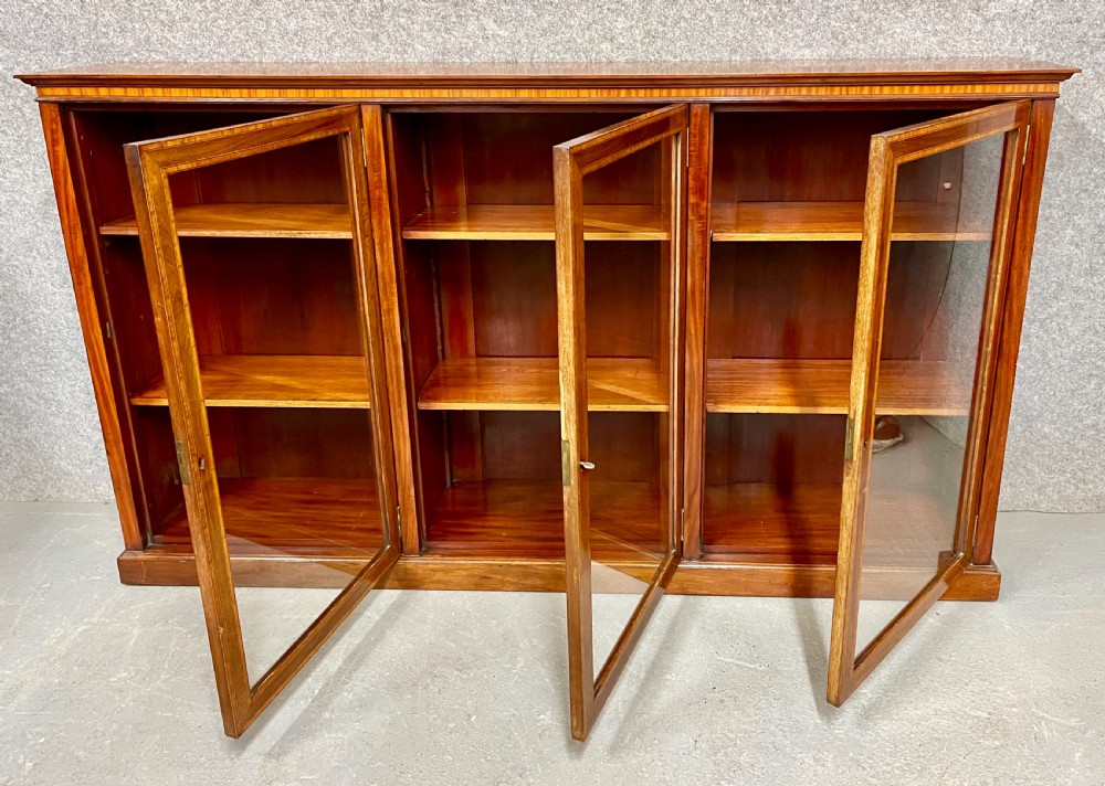 A wide, low Edwardian mahogany bookcase featuring three glass doors accented with satinwood crossbanding and internal wooden shelves on a plinth base. View VI