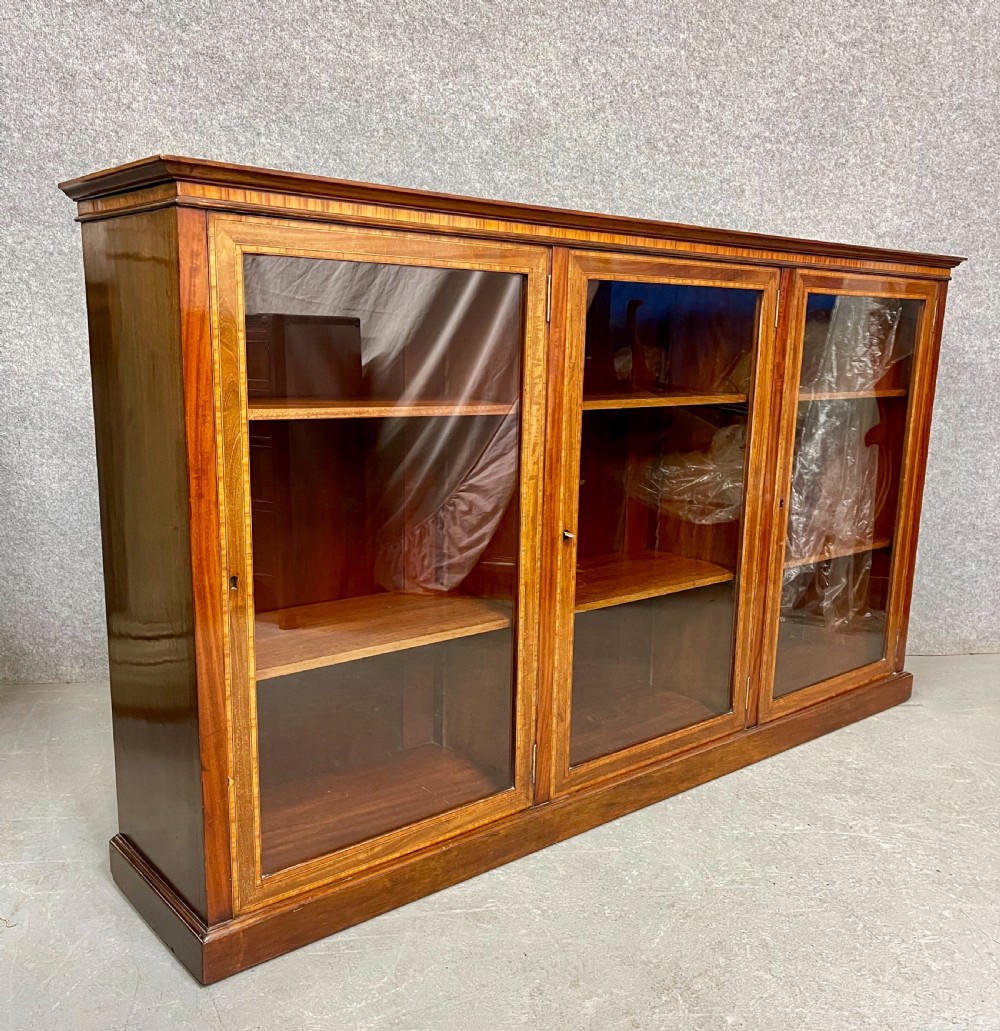 A wide, low Edwardian mahogany bookcase featuring three glass doors accented with satinwood crossbanding and internal wooden shelves on a plinth base.