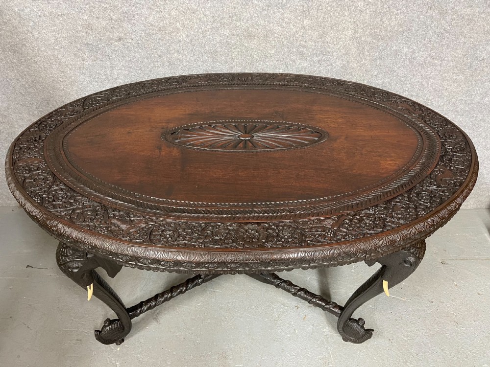 An oval Anglo-Indian dark wood centre table featuring an intricately carved floral border, a central radiating oval medallion, and legs sculpted as elephant heads with white tusks. View X