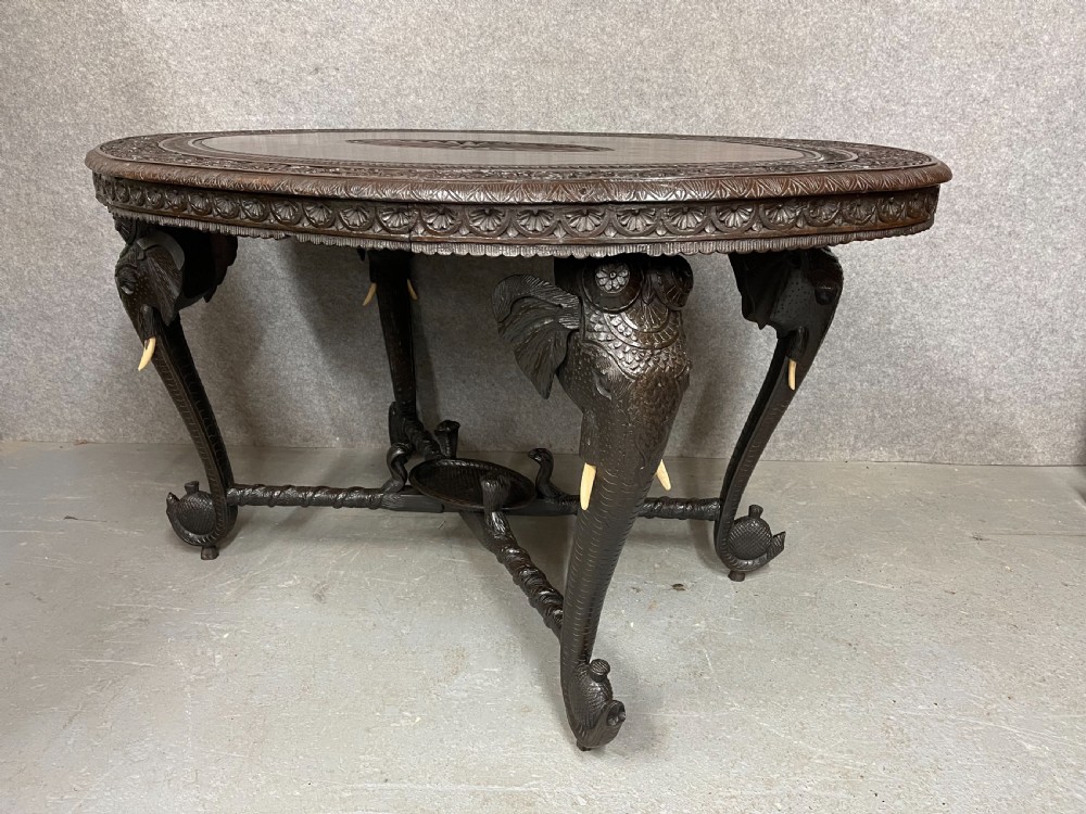 An oval Anglo-Indian dark wood centre table featuring an intricately carved floral border, a central radiating oval medallion, and legs sculpted as elephant heads with white tusks. View II
