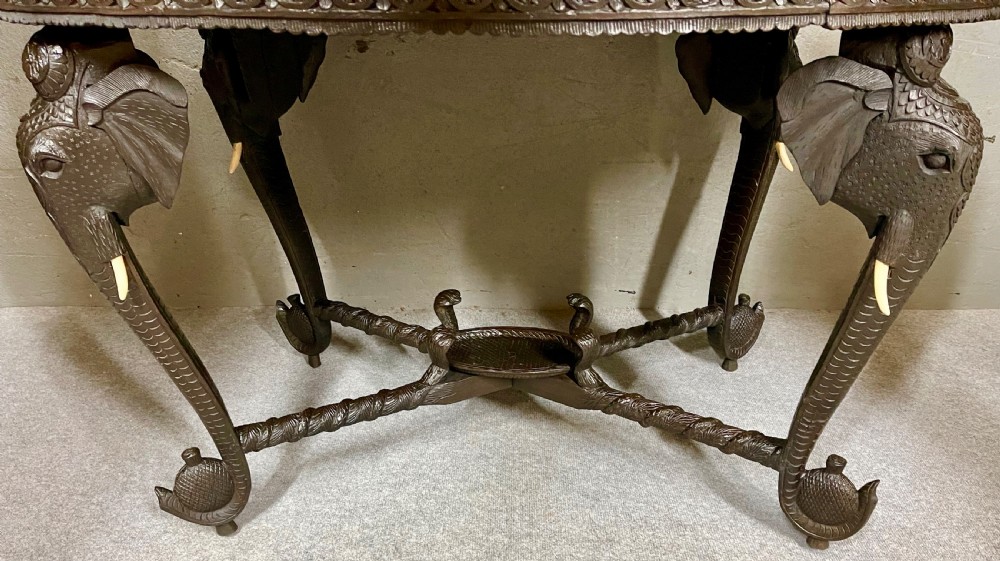 An oval Anglo-Indian dark wood centre table featuring an intricately carved floral border, a central radiating oval medallion, and legs sculpted as elephant heads with white tusks. View VI