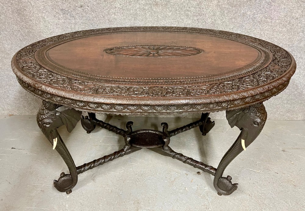 An oval Anglo-Indian dark wood centre table featuring an intricately carved floral border, a central radiating oval medallion, and legs sculpted as elephant heads with white tusks.