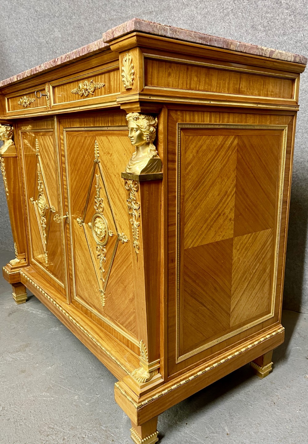 A pair of French Neoclassical side cabinets featuring satinwood parquetry, intricate gilt-bronze mounts, corner bust sculptures, and pink marble tops. View VIII