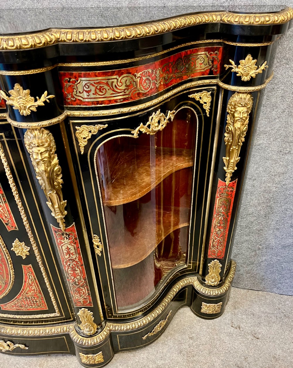 An ebonised serpentine side cabinet featuring red tortoiseshell and brass Boulle marquetry, gilt-bronze ormolu mounts, a central oval-panelled door, and two curved side glazed display sections. View IV