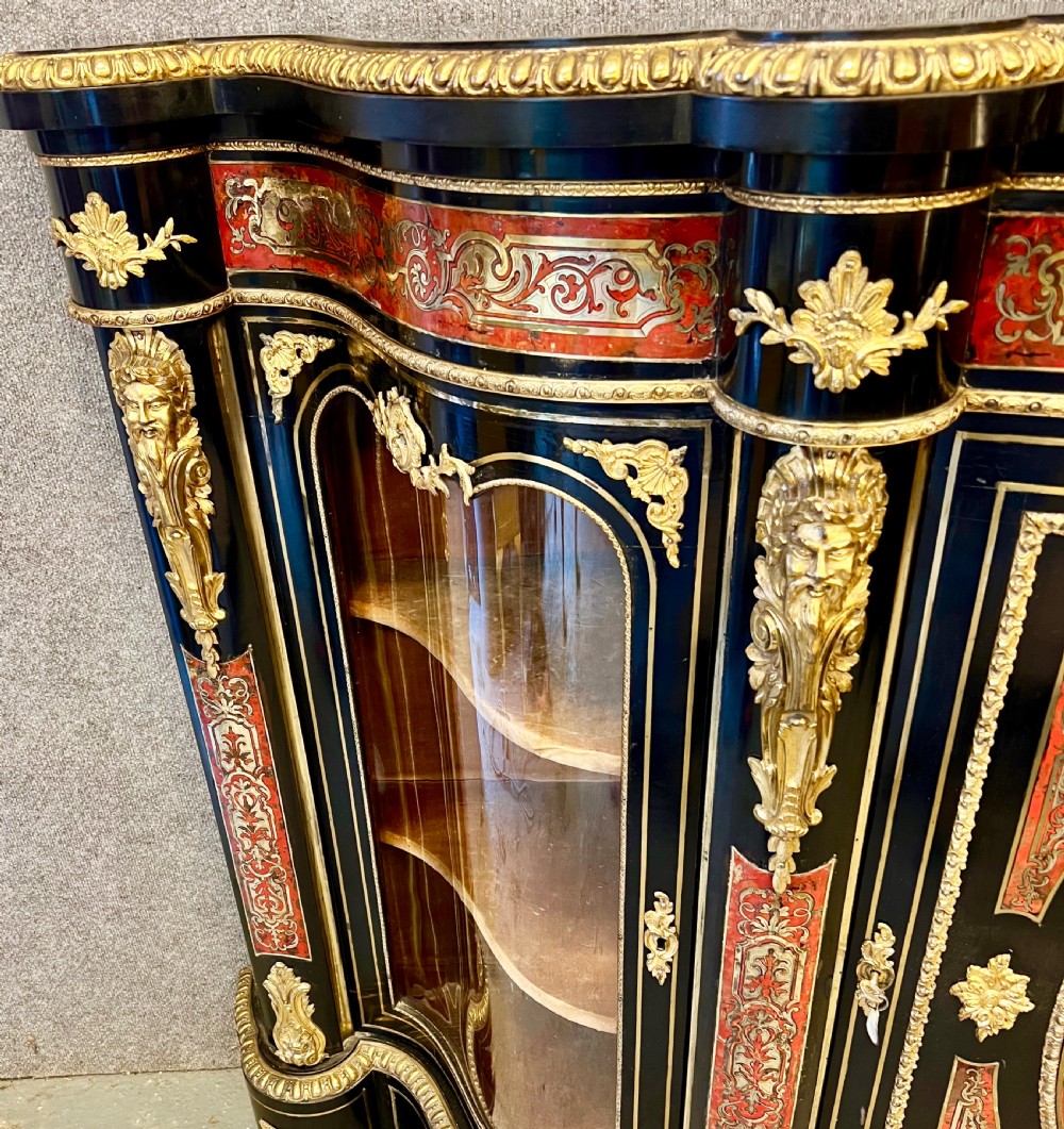 An ebonised serpentine side cabinet featuring red tortoiseshell and brass Boulle marquetry, gilt-bronze ormolu mounts, a central oval-panelled door, and two curved side glazed display sections. View V