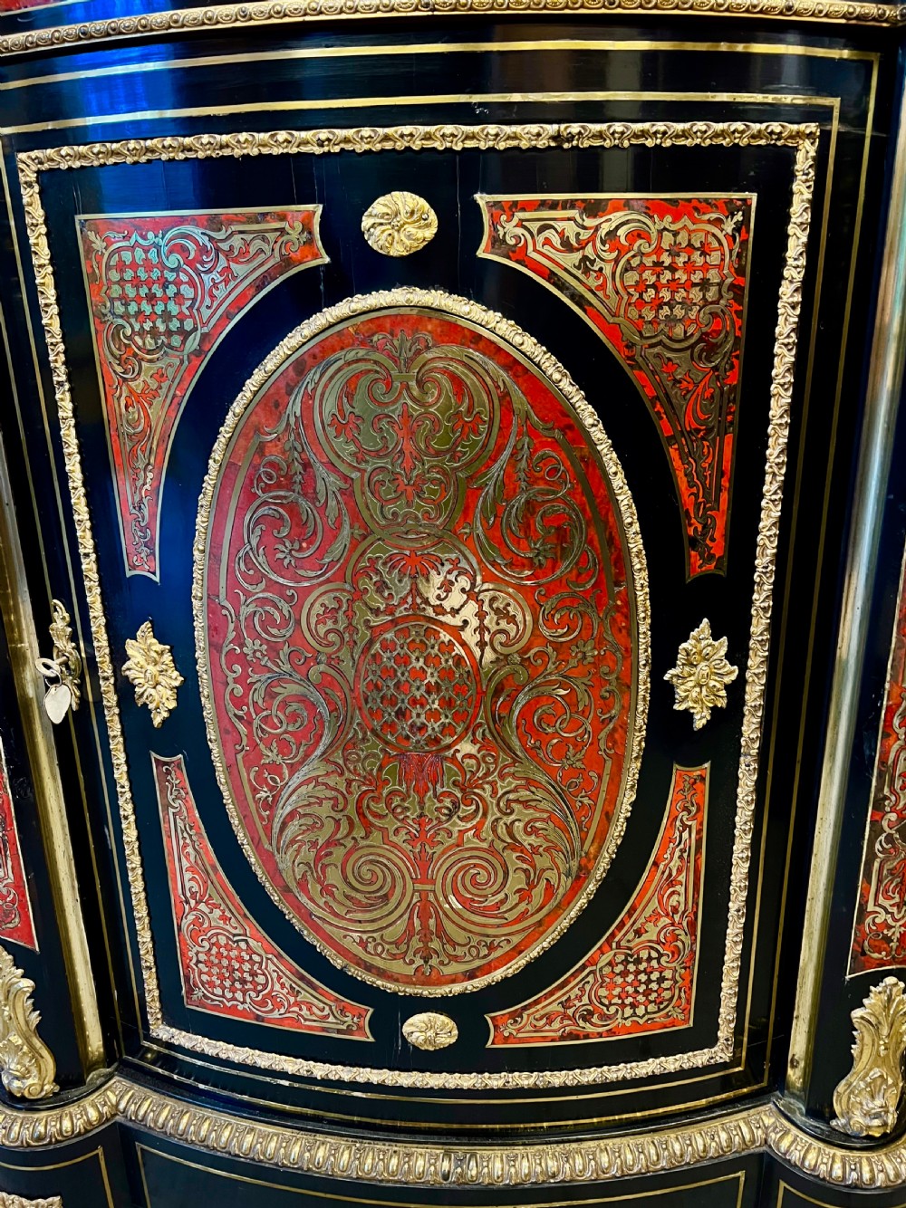 An ebonised serpentine side cabinet featuring red tortoiseshell and brass Boulle marquetry, gilt-bronze ormolu mounts, a central oval-panelled door, and two curved side glazed display sections. View VIII
