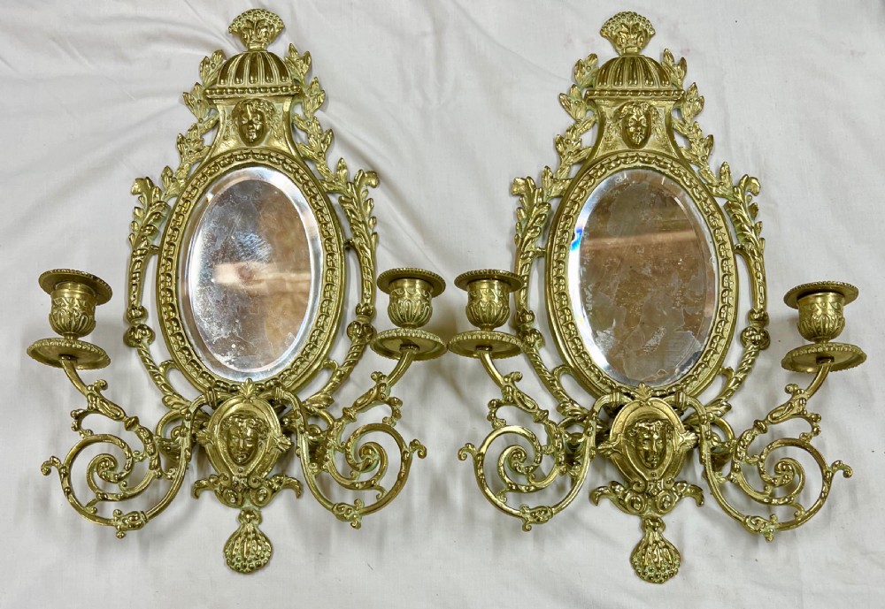 A pair of gilt brass girandole wall mirrors with oval bevelled glass plates, classical mask motifs, acanthus scrolls, and twin candle holders.