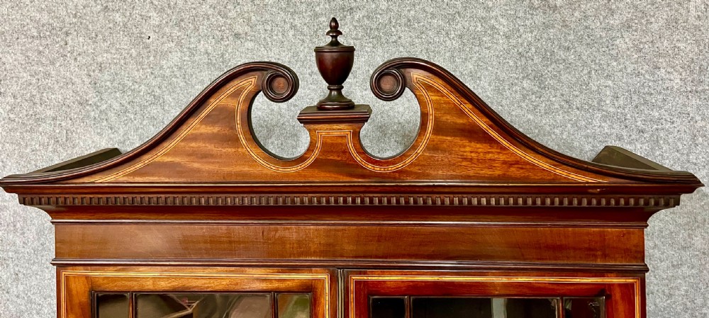 A Sheraton Revival mahogany cabinet featuring a swan-neck pediment, glazed upper doors with geometric bars, inlaid satinwood stringing, and multiple drawers set on tapered square legs. View VI