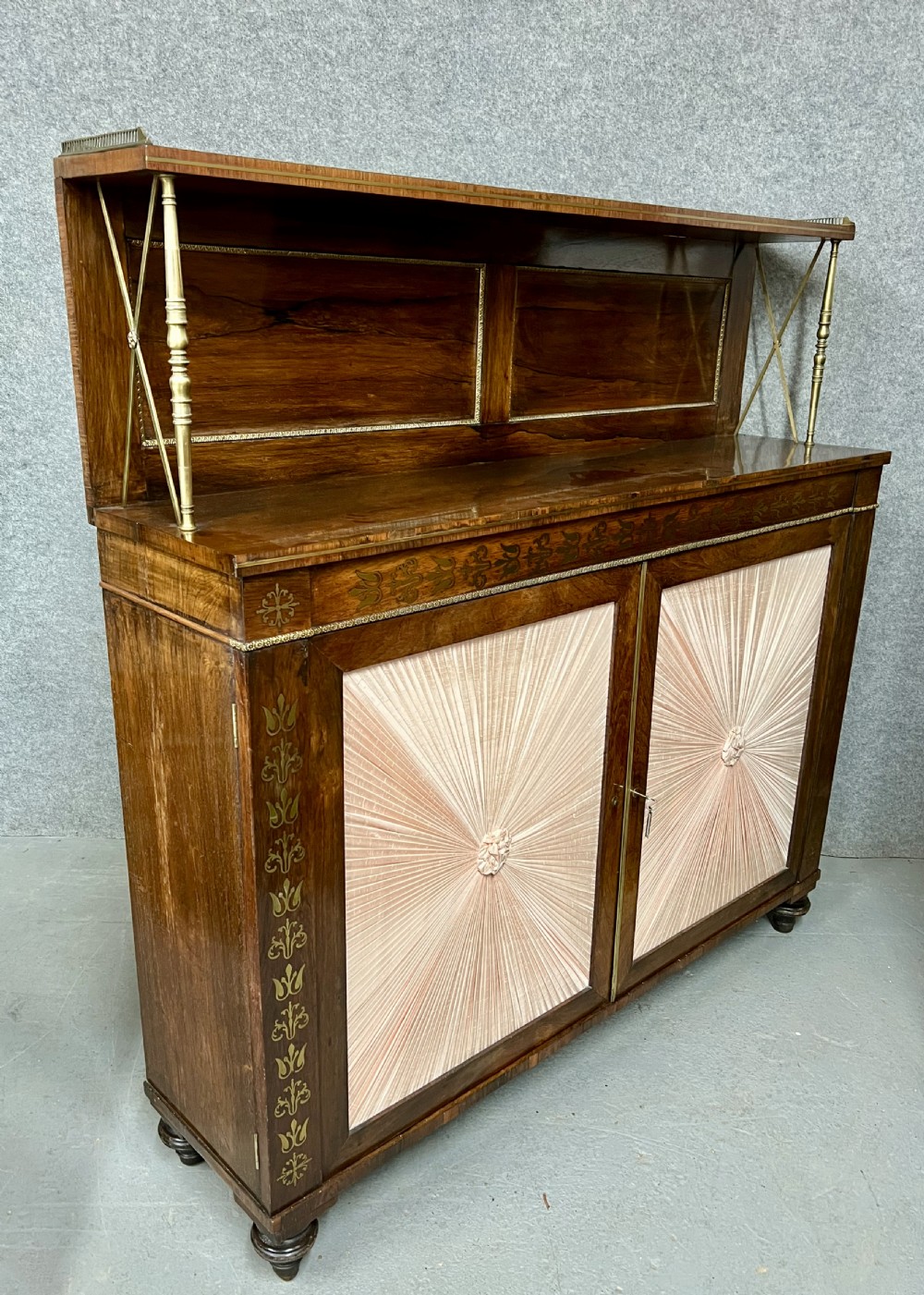 A Regency rosewood chiffonier featuring ornate brass marquetry inlay, twin doors with pleated fabric panels, and a raised top shelf supported by brass columns.