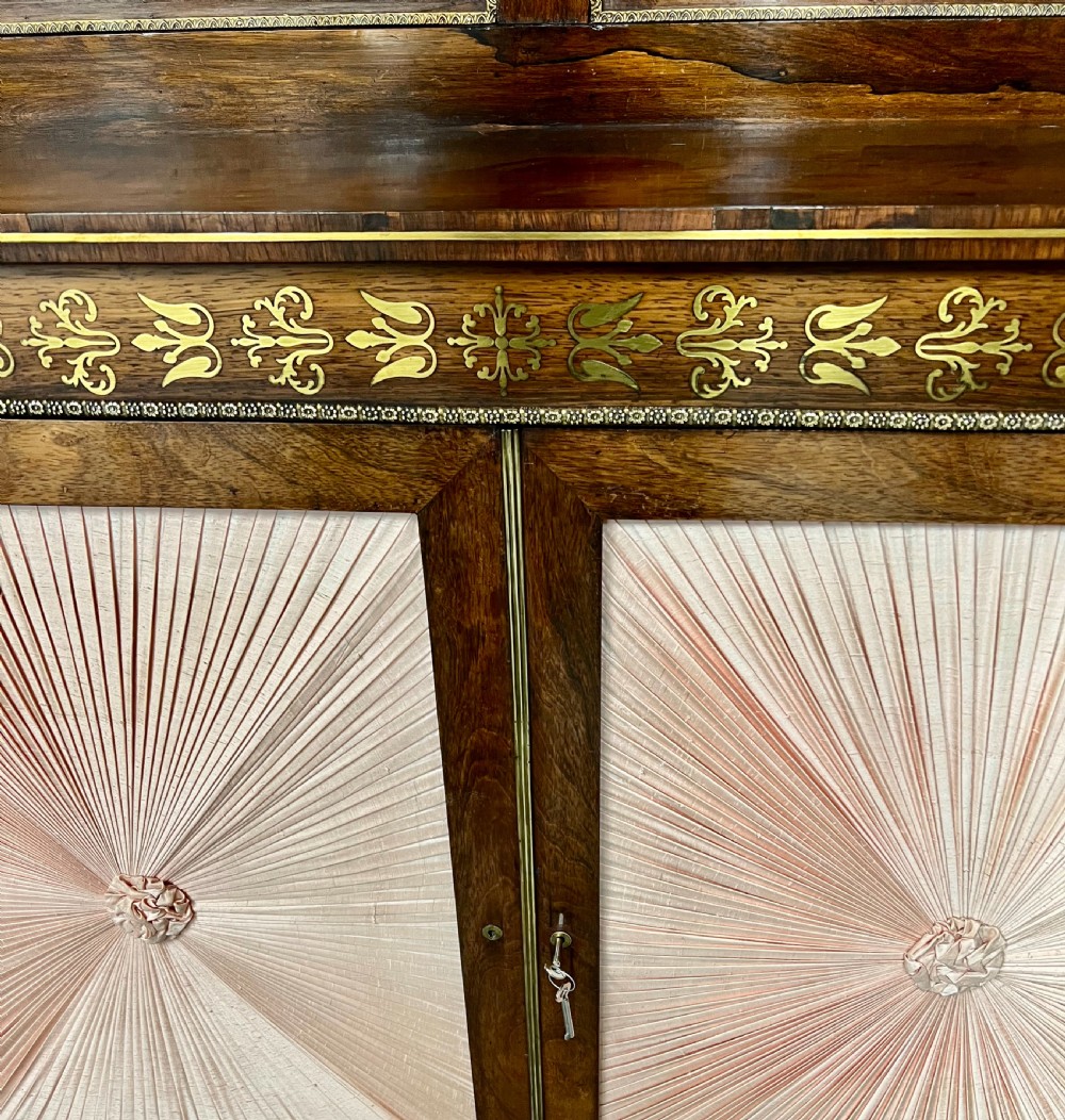 A Regency rosewood chiffonier featuring ornate brass marquetry inlay, twin doors with pleated fabric panels, and a raised top shelf supported by brass columns. View IV