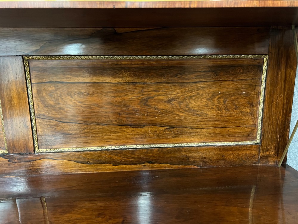 A Regency rosewood chiffonier featuring ornate brass marquetry inlay, twin doors with pleated fabric panels, and a raised top shelf supported by brass columns. View X
