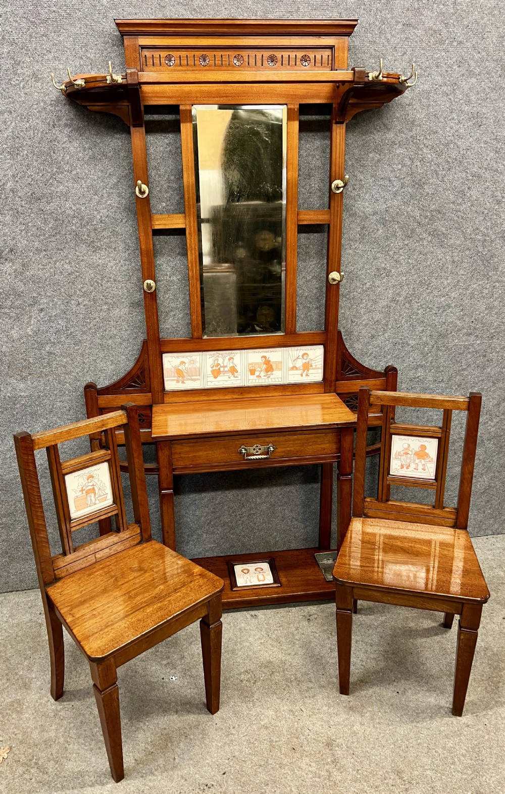 A late 19th-century mahogany hall stand featuring a central mirror, brass hooks, inlaid Minton ceramic tiles, and two matching side chairs.
