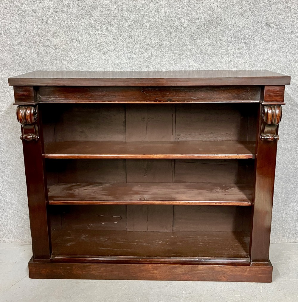 A low Victorian dark mahogany open bookcase featuring three horizontal shelves, carved decorative corbels on the upper corners, and a solid plinth base.