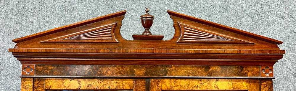 A Victorian burr walnut bookcase cabinet featuring a broken pediment with a central urn finial, glazed upper doors, two drawers, and carved lower cupboard doors. View V