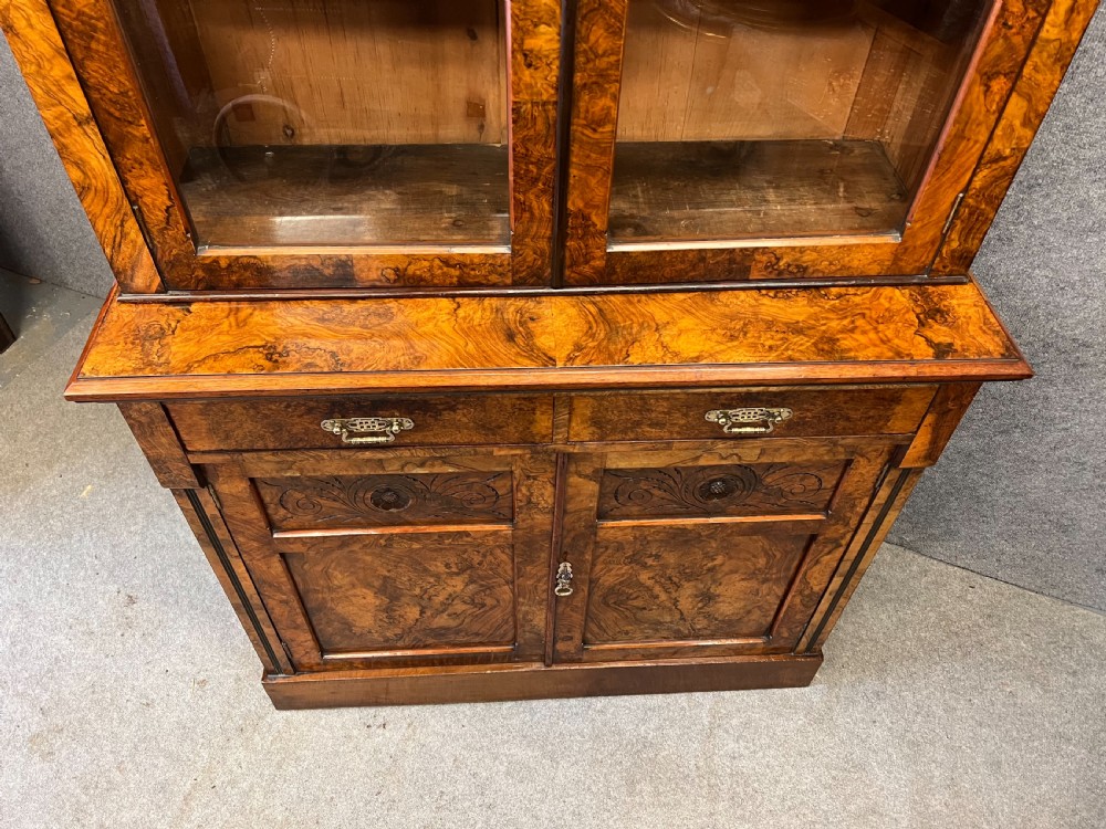 A Victorian burr walnut bookcase cabinet featuring a broken pediment with a central urn finial, glazed upper doors, two drawers, and carved lower cupboard doors. View VII