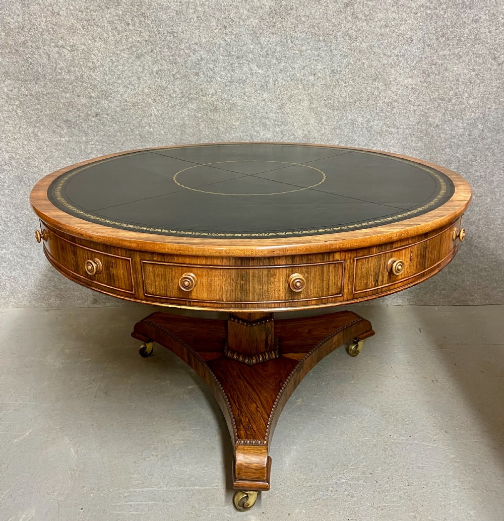 A Regency period circular rosewood drum table featuring a black gold-tooled leather writing top, frieze drawers, and a tripartite pedestal base on brass castors.