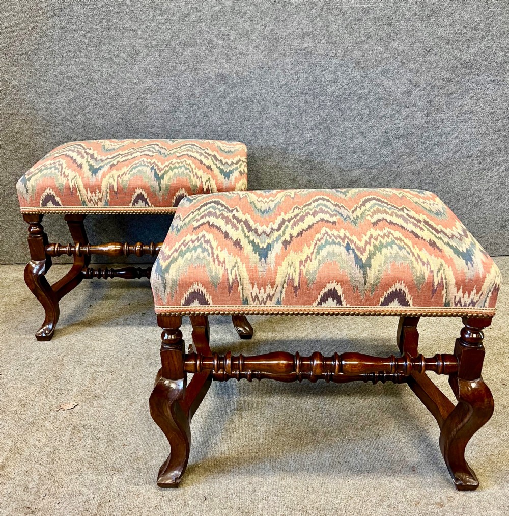 A pair of 18th-century style walnut stools featuring turned stretchers, curved legs, and patterned flame-stitch upholstery finished with brass studs.