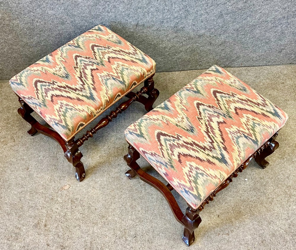 A pair of 18th-century style walnut stools featuring turned stretchers, curved legs, and patterned flame-stitch upholstery finished with brass studs. View V