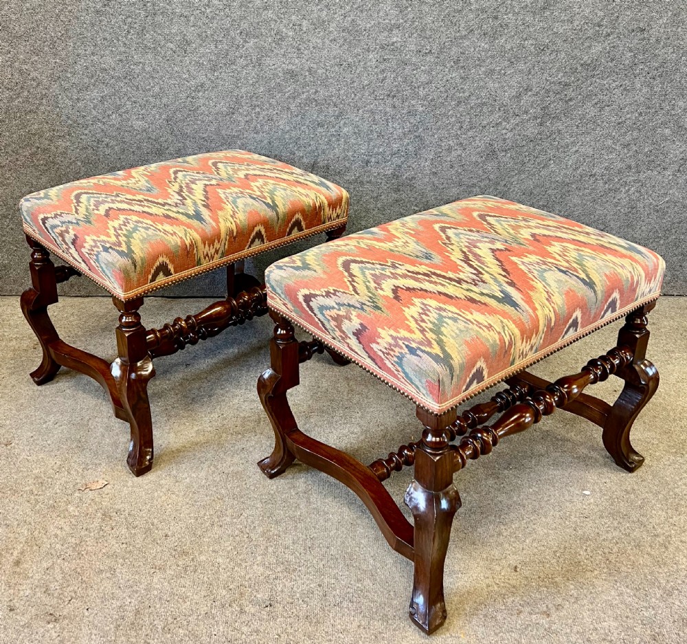 A pair of 18th-century style walnut stools featuring turned stretchers, curved legs, and patterned flame-stitch upholstery finished with brass studs. View VI