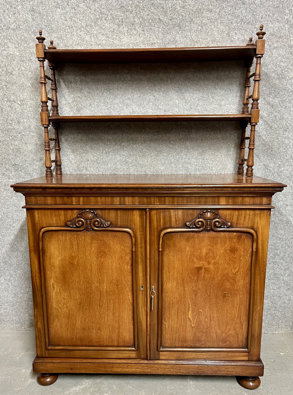 An antique mahogany chiffonier featuring two panelled cupboard doors with carved shell details below a tiered open bookcase supported by turned spindles.
