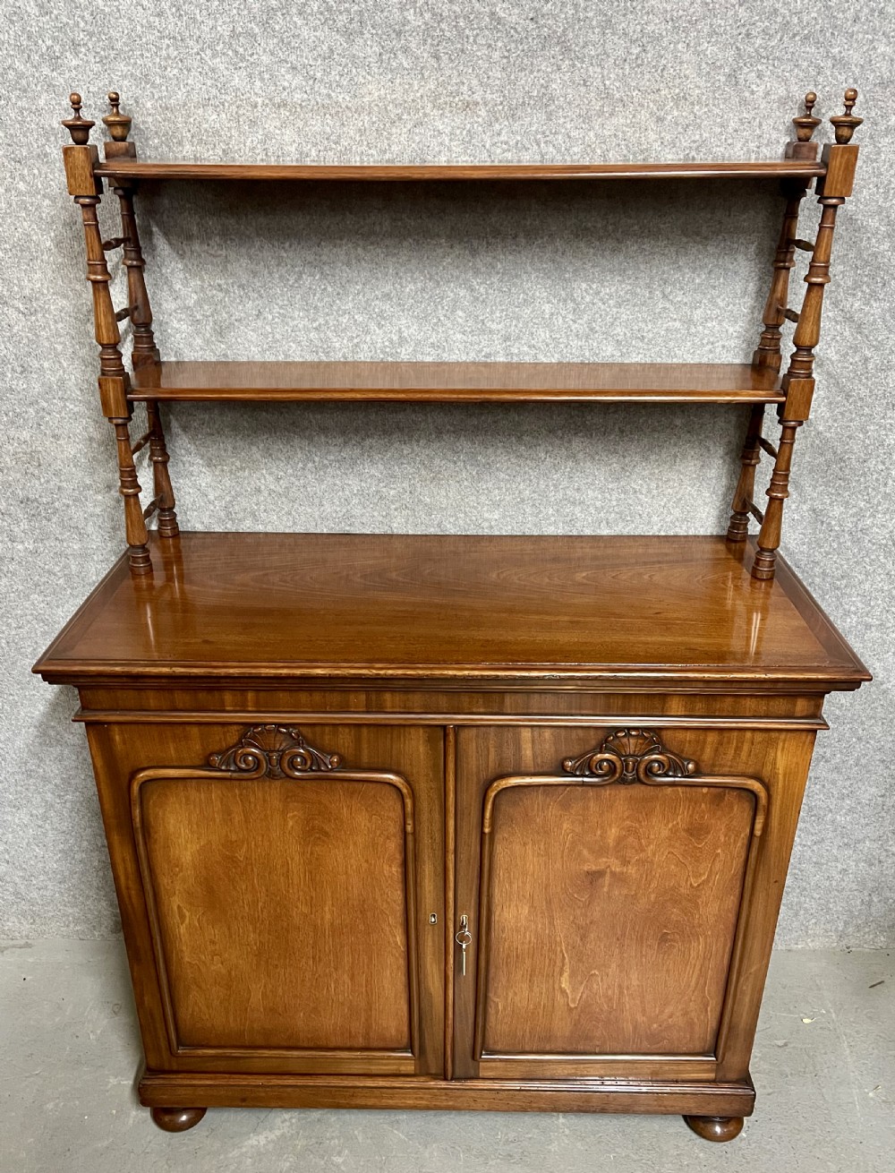 An antique mahogany chiffonier featuring two panelled cupboard doors with carved shell details below a tiered open bookcase supported by turned spindles. View VI