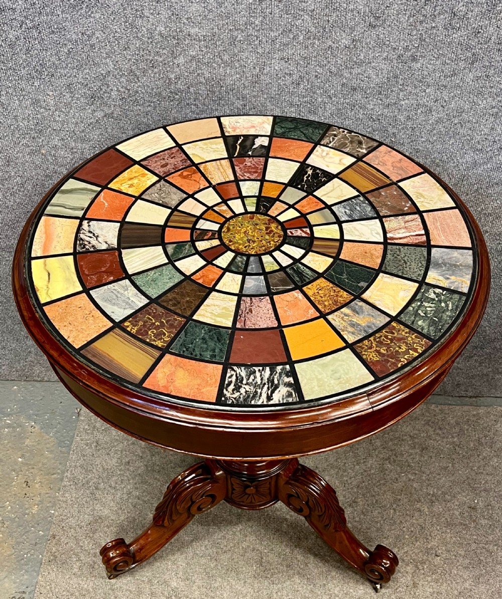A circular Grand Tour style table featuring a radiating geometric mosaic top of colourful specimen marble inlays supported on a carved mahogany pedestal base with scrolled feet.