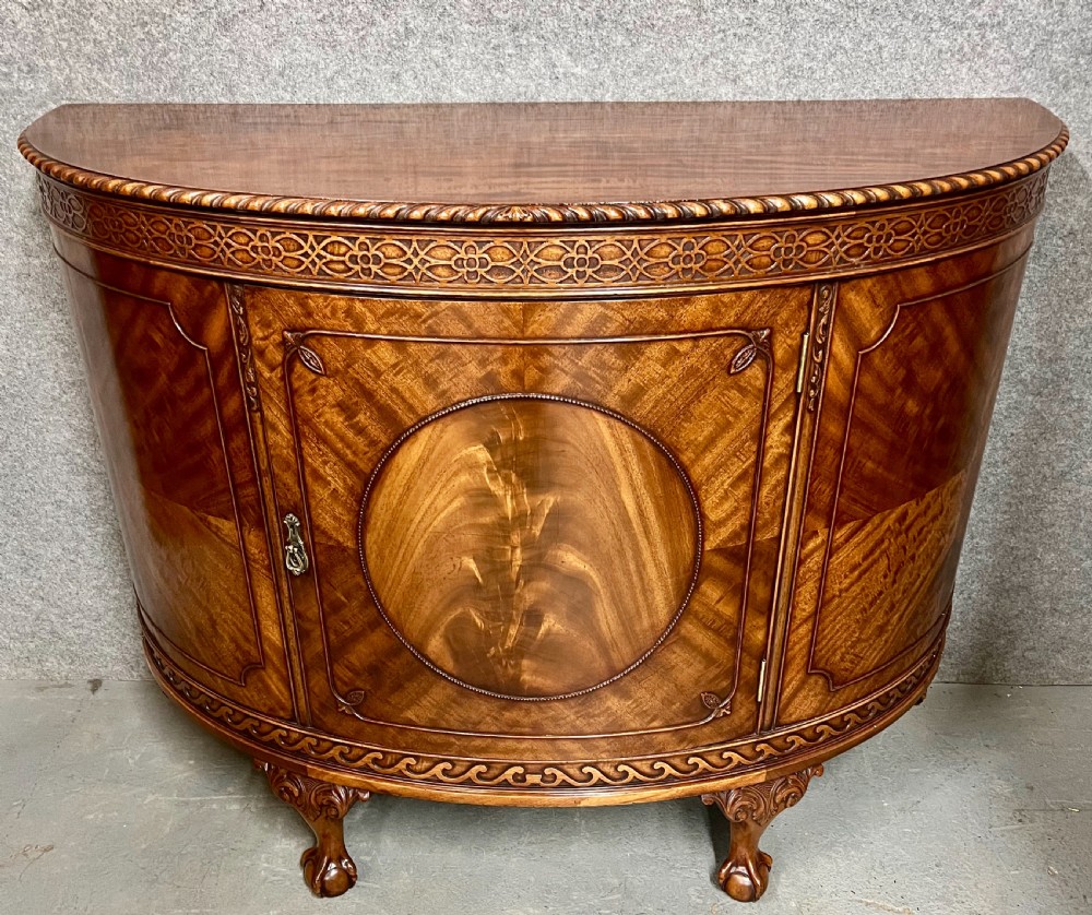 A semi-circular Chippendale style mahogany demi-lune side cabinet featuring flame mahogany veneers, a carved border, a central oval door panel, and carved claw and ball feet.