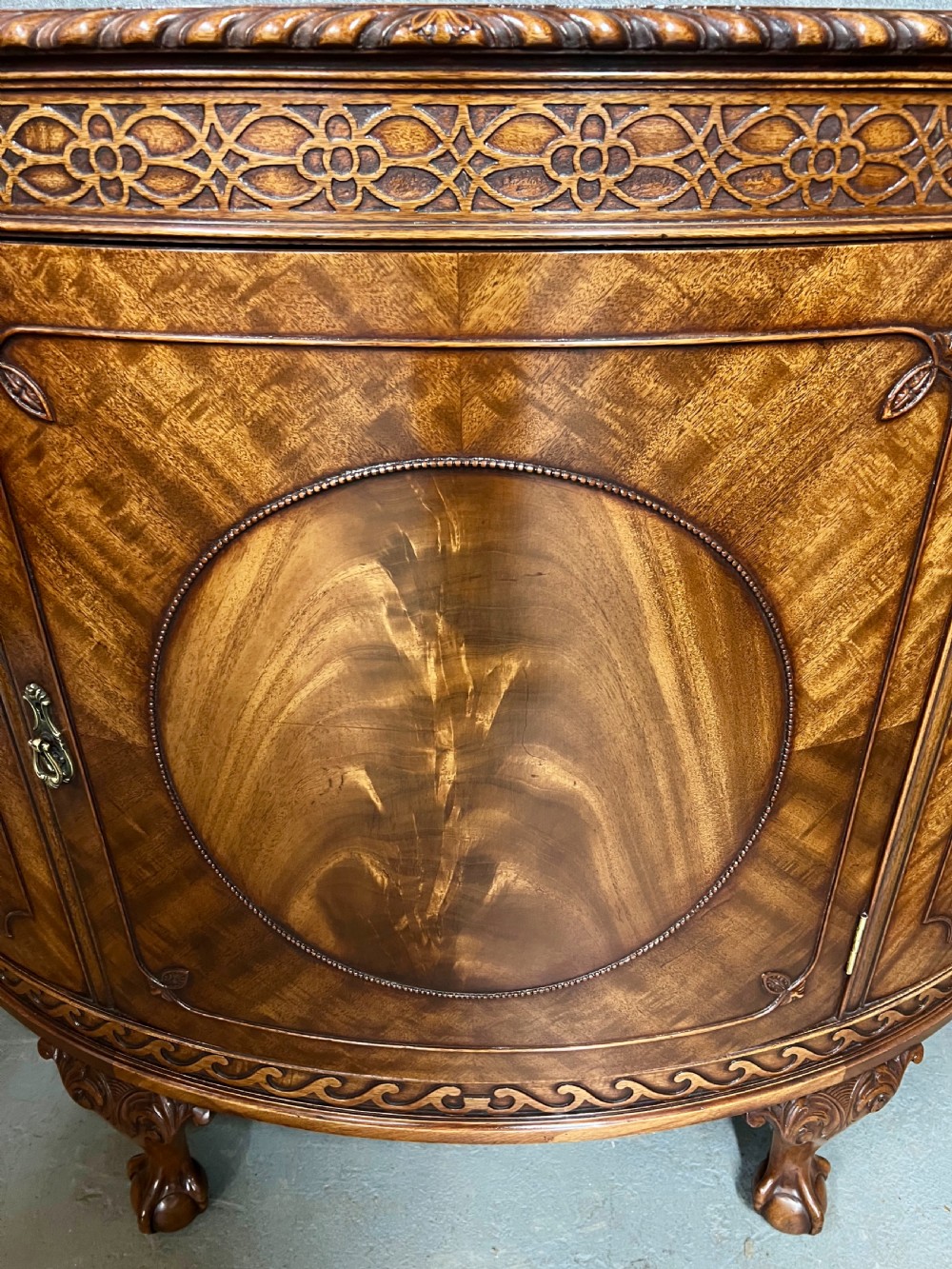 A semi-circular Chippendale style mahogany demi-lune side cabinet featuring flame mahogany veneers, a carved border, a central oval door panel, and carved claw and ball feet. View IV