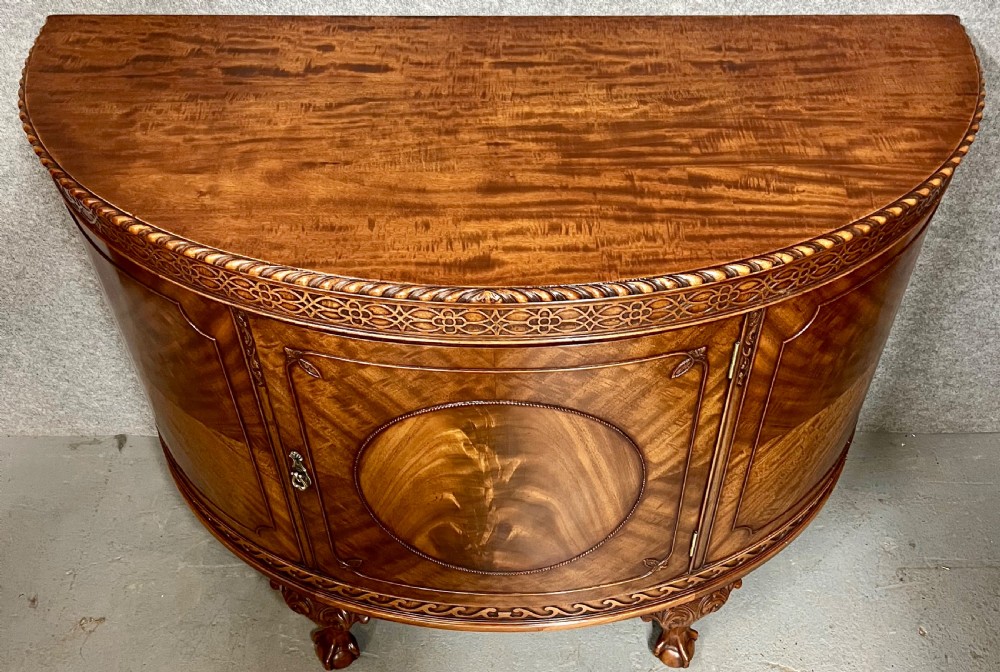 A semi-circular Chippendale style mahogany demi-lune side cabinet featuring flame mahogany veneers, a carved border, a central oval door panel, and carved claw and ball feet. View VIII