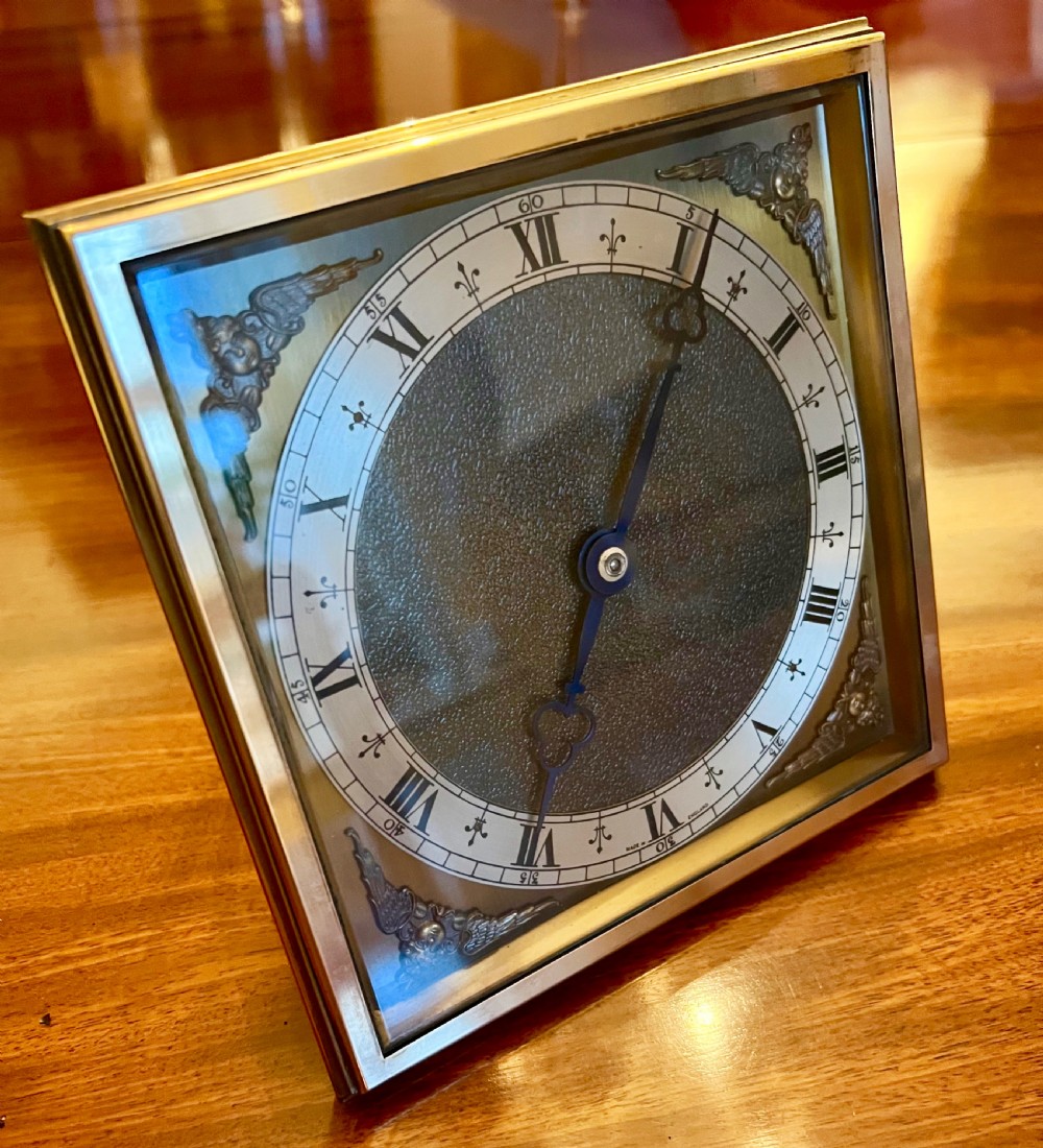 A square brass framed desk clock featuring a silvered chapter ring with Roman numerals, blued steel hands, and corner cherub spandrel accents. View II