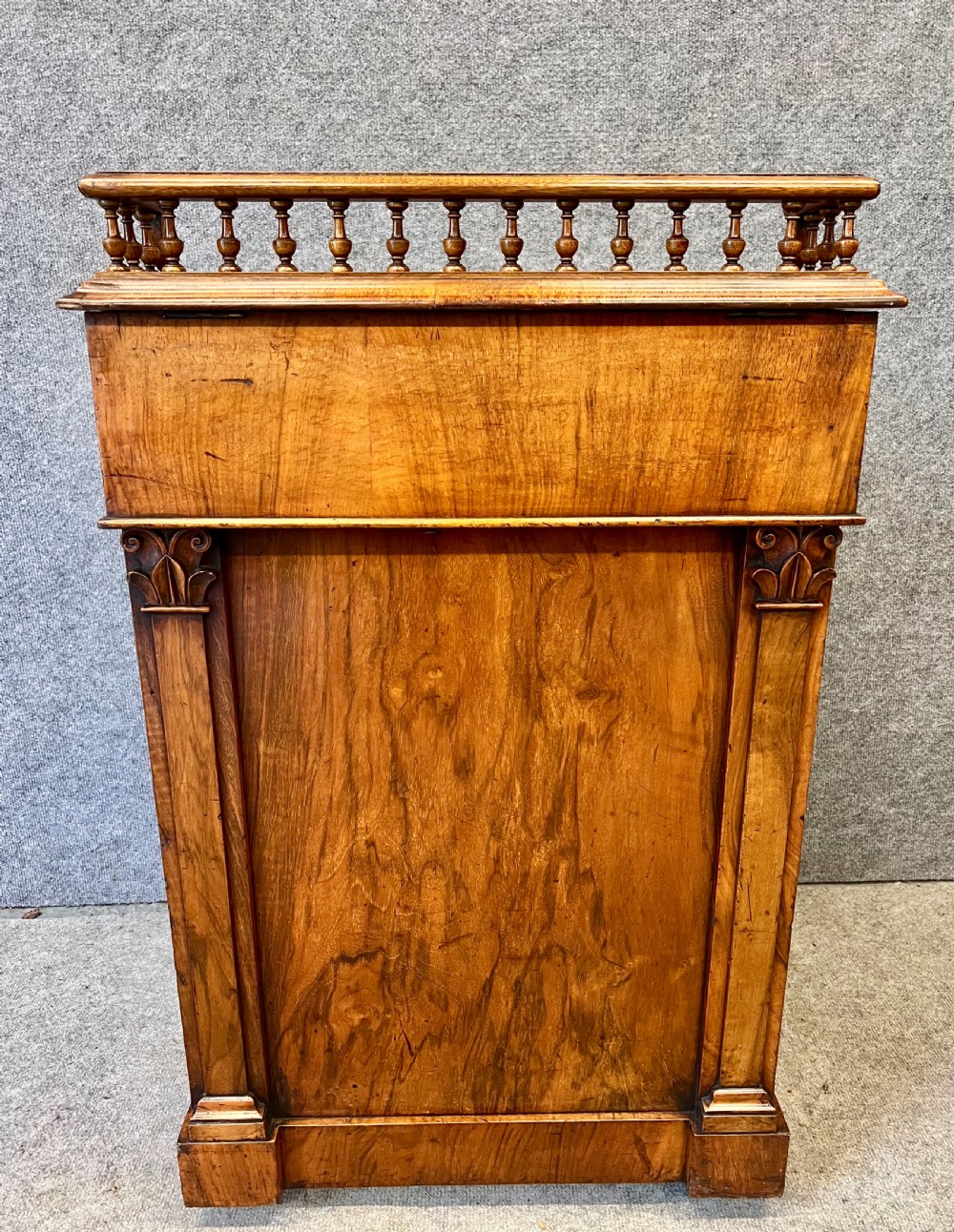 A Victorian walnut slide-top Davenport desk featuring a spindle gallery rail, leather writing surface, side drawers with turned knobs, and carved scroll supports. View VI