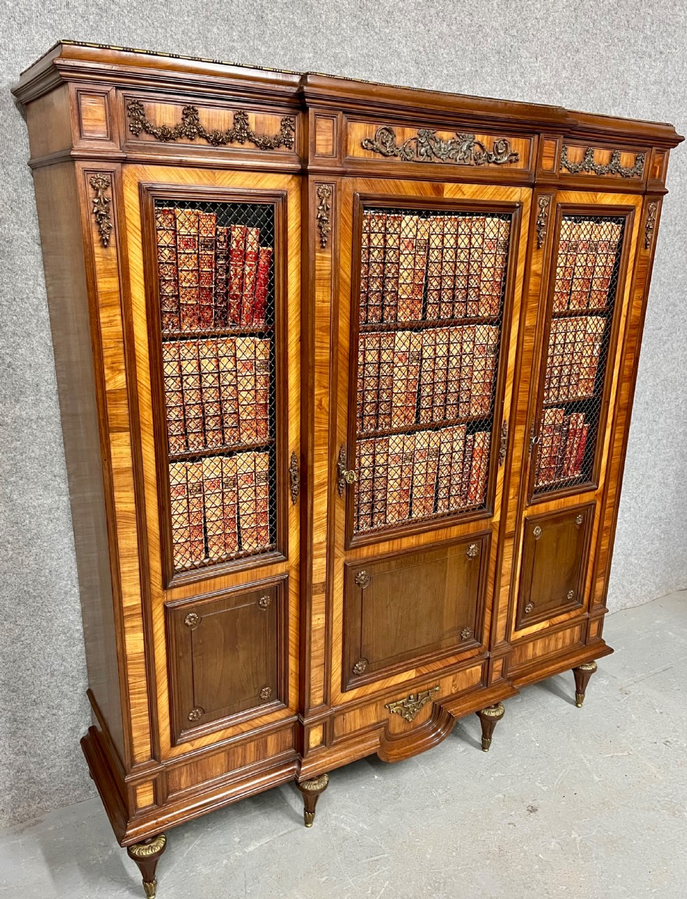 An antique French three-door bookcase crafted from kingwood and mahogany, featuring wire-mesh upper doors, lower timber panel doors, turned legs, and carved decorative crests. View III