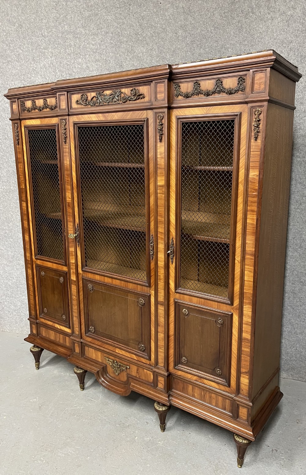 An antique French three-door bookcase crafted from kingwood and mahogany, featuring wire-mesh upper doors, lower timber panel doors, turned legs, and carved decorative crests. View IV
