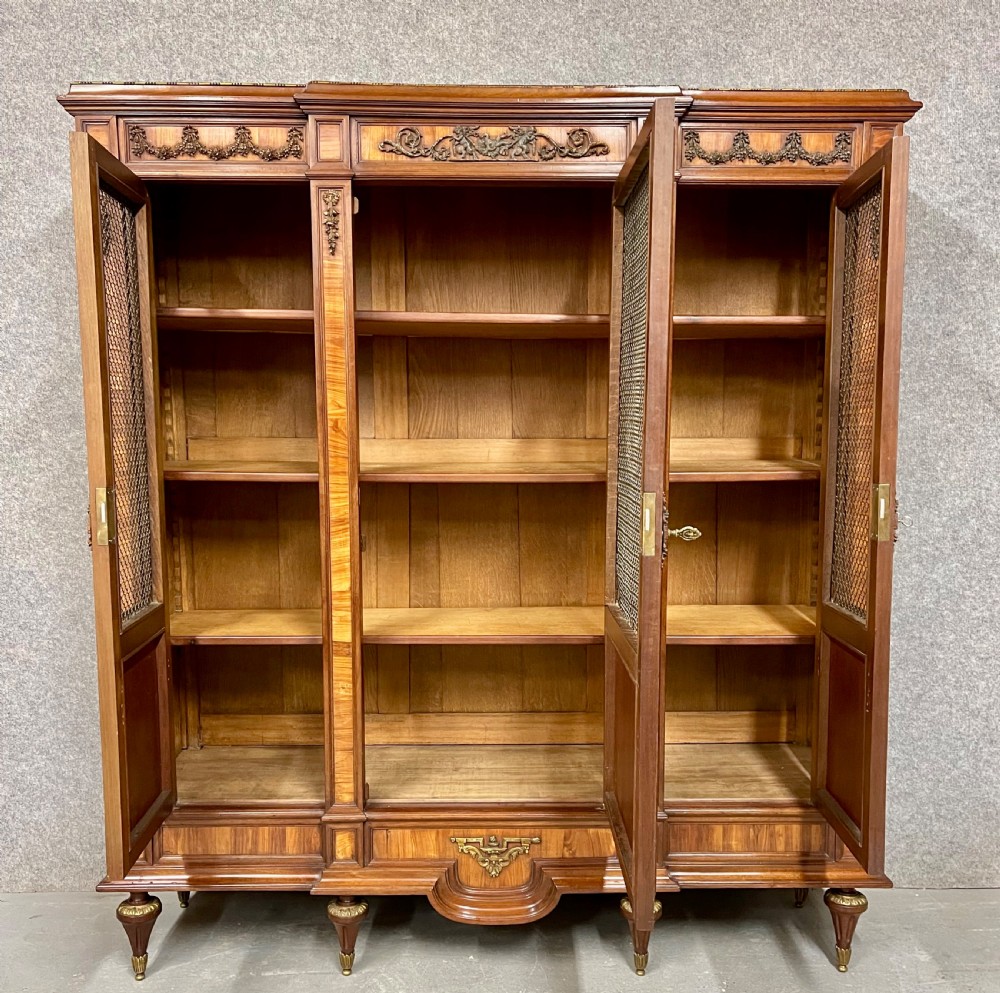 An antique French three-door bookcase crafted from kingwood and mahogany, featuring wire-mesh upper doors, lower timber panel doors, turned legs, and carved decorative crests. View VI